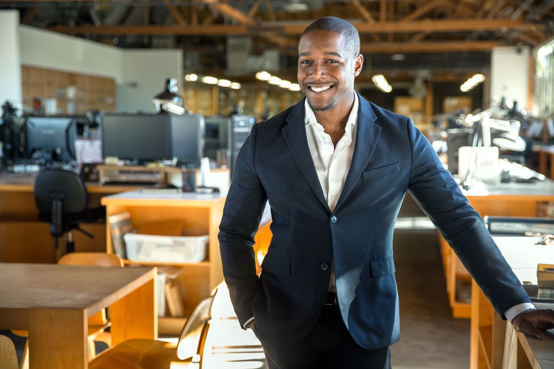 Smiling man in a navy blazer standing in a modern office with desks and computers.