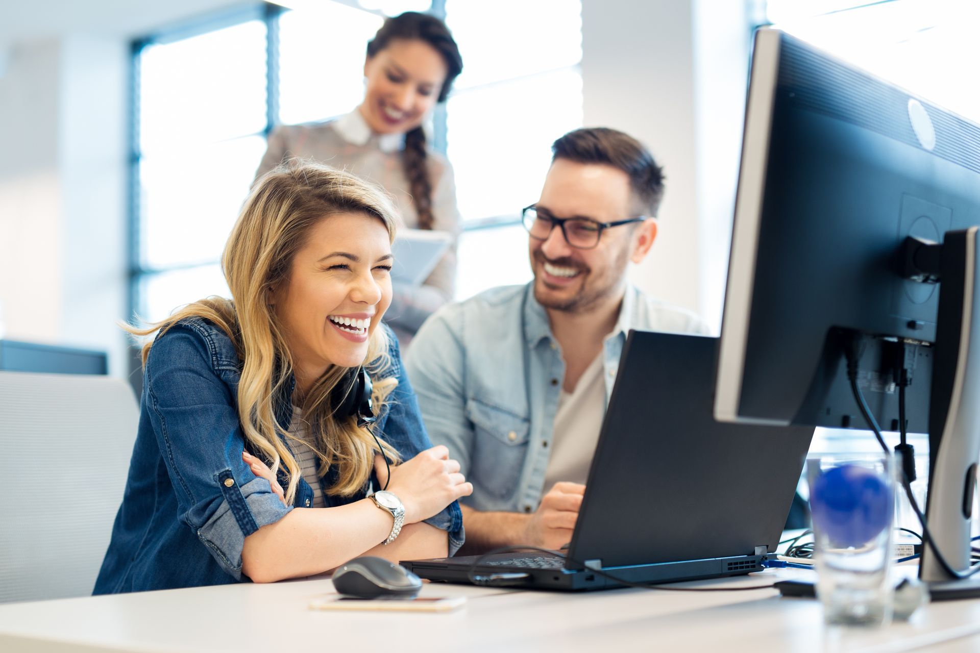 Colleagues smiling at a desktop computer in a bright office, with a laptop on the desk
