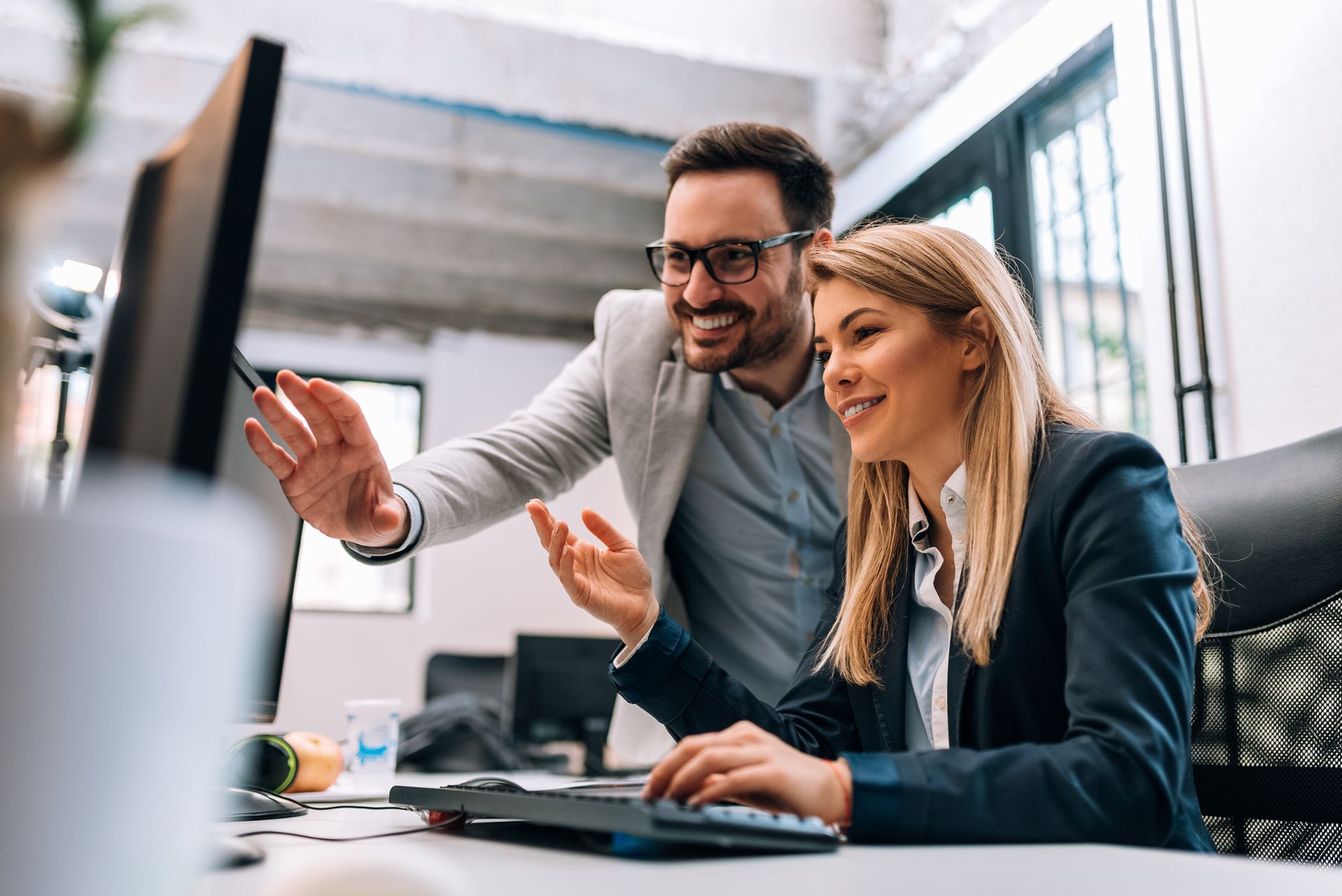 Two coworkers smiling and collaborating at a desk, with one pointing at a computer screen