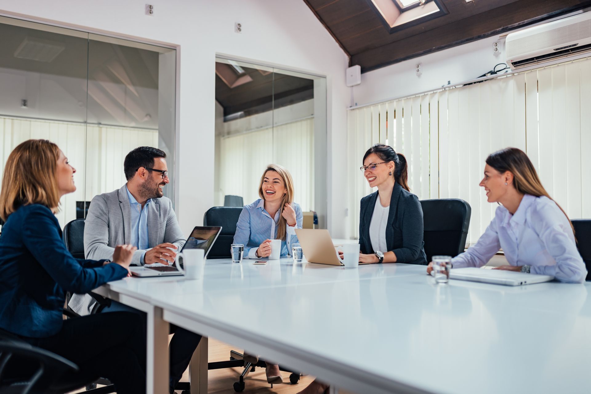 People in a bright conference room discussing around a white table with laptops and papers