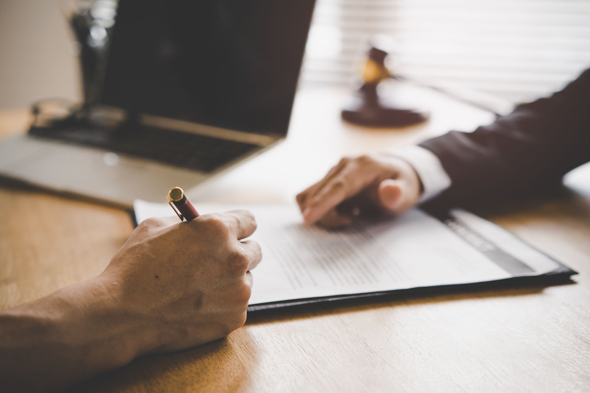 Two people sign documents at a wooden desk near a laptop in warm office light