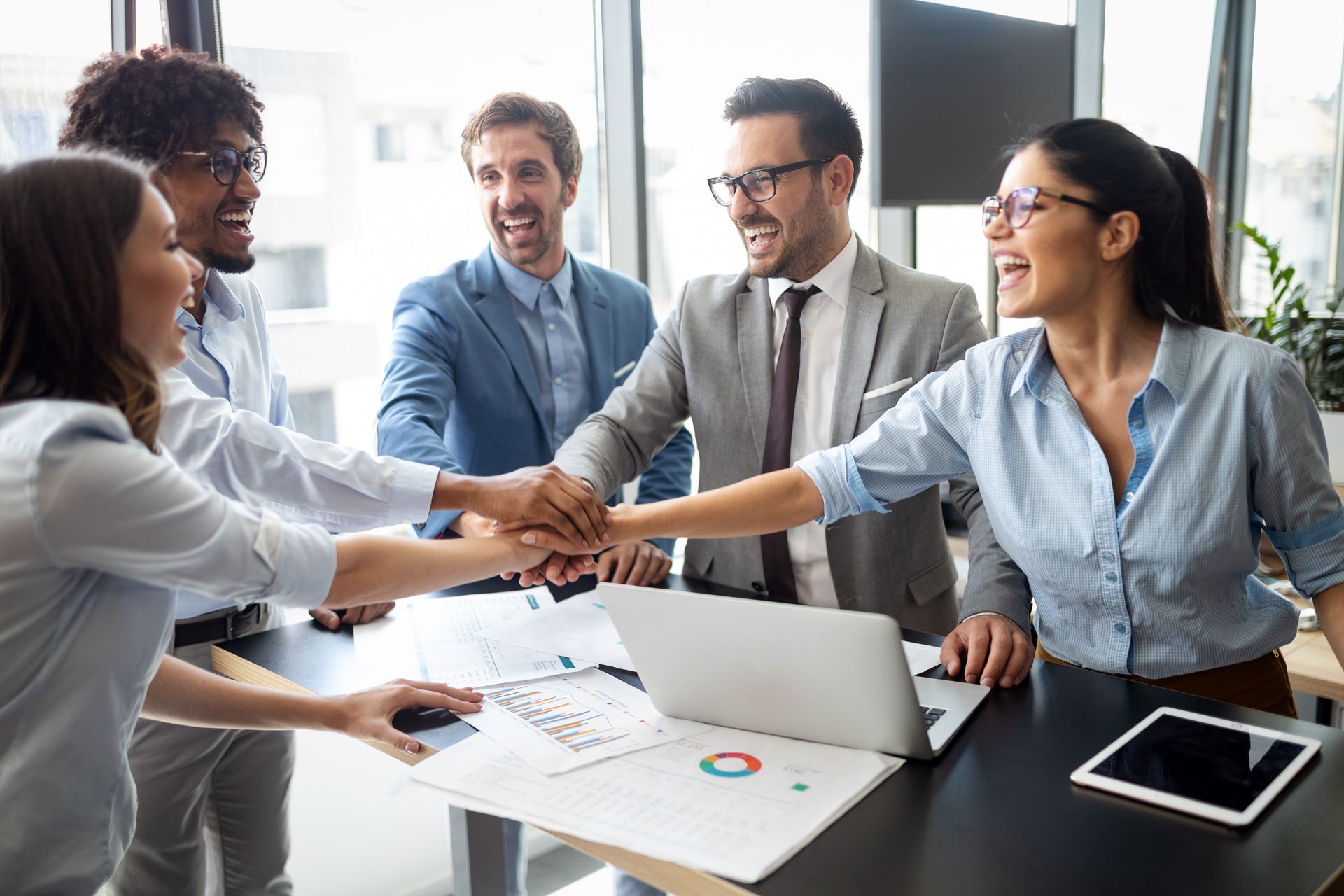 Team members in a meeting, smiling and shaking hands across a table with a laptop and papers.