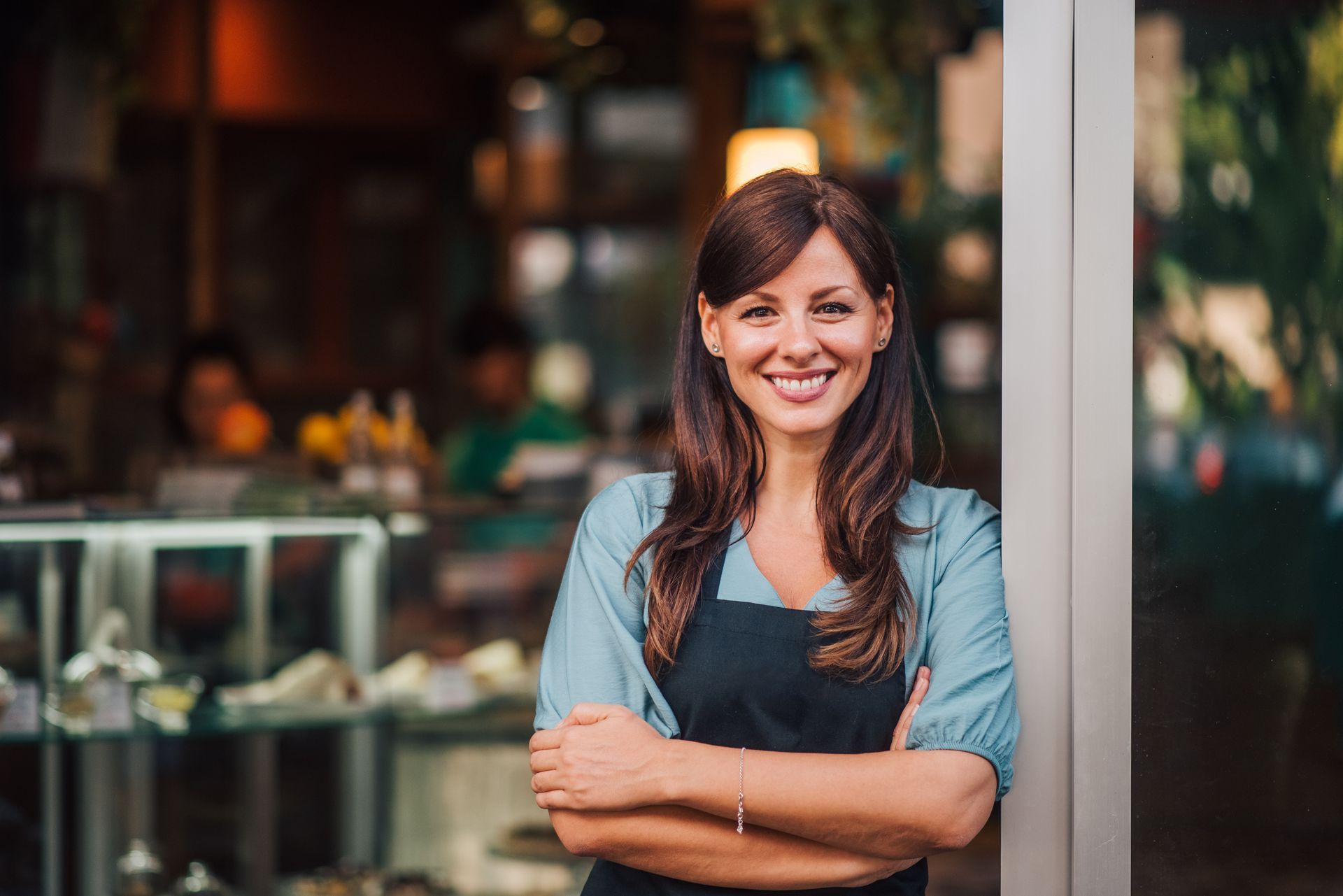 Smiling cafe worker in a dark apron, standing with arms crossed near a counter.