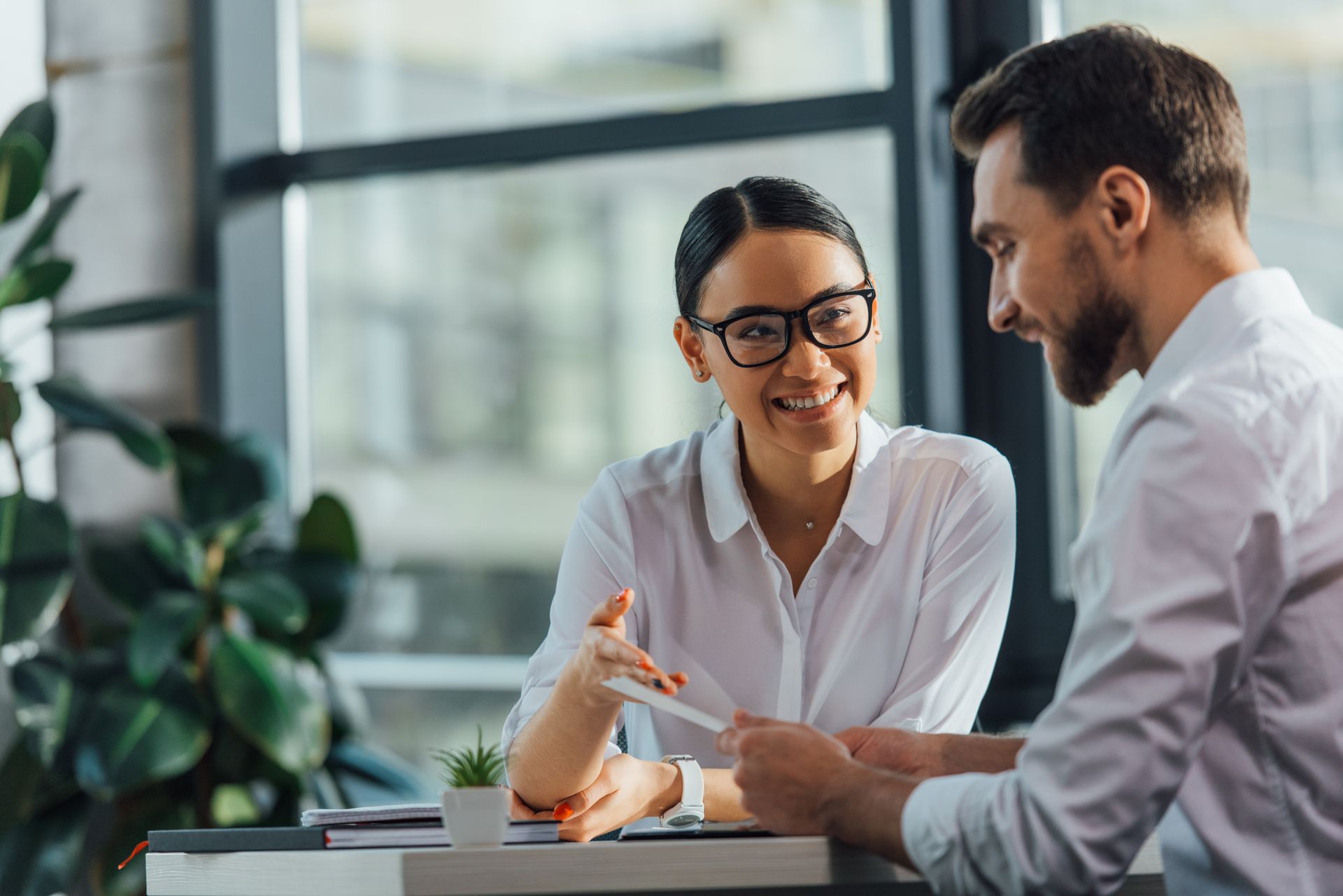 Two coworkers in a bright office discuss documents at a desk, one smiling and pointing.