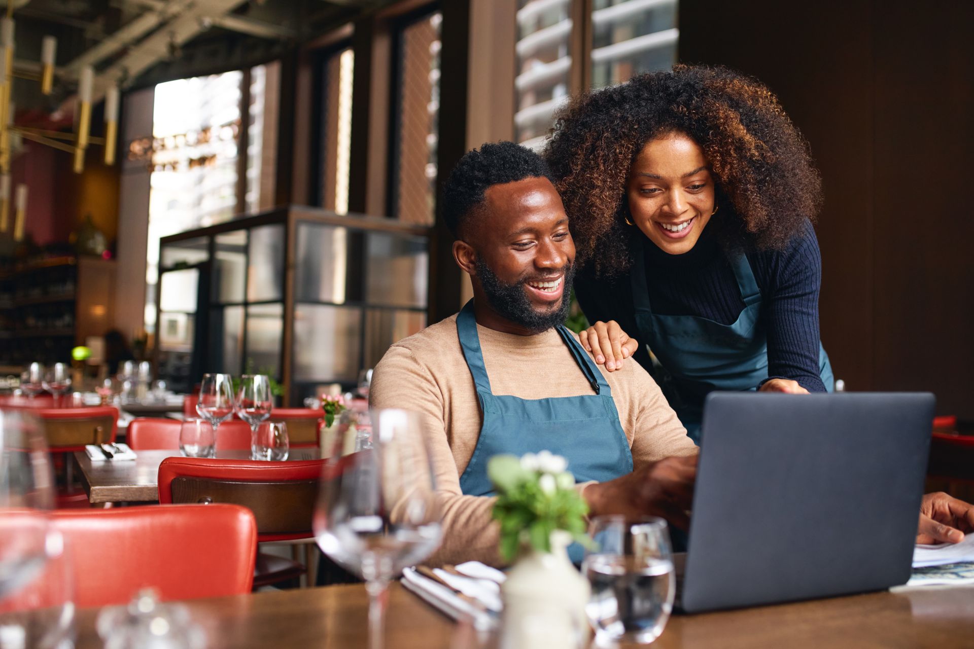 Two people smiling at a laptop in a cozy café with red chairs and wine glasses on the table