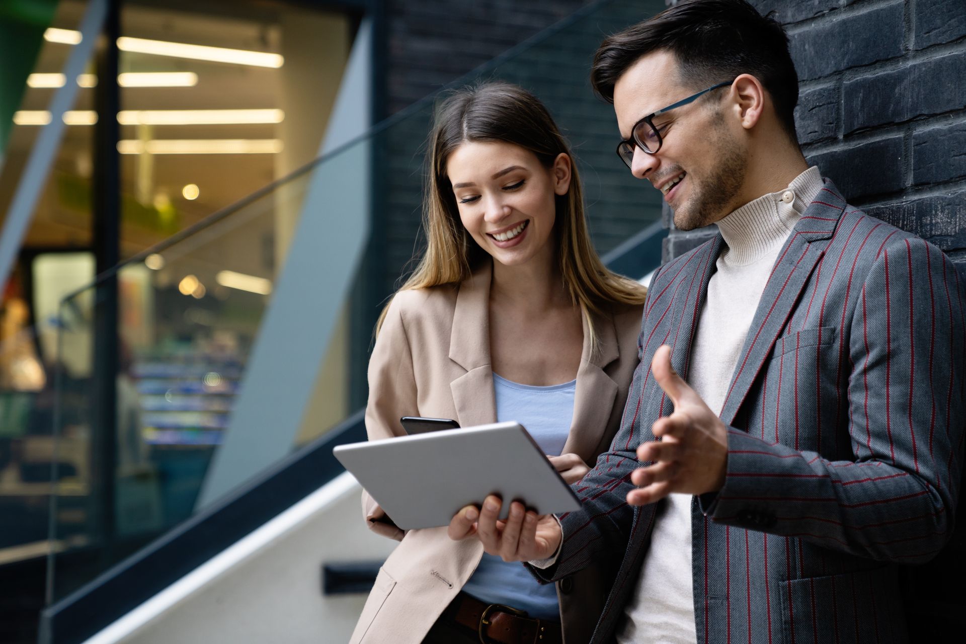 Two coworkers reviewing a tablet and smiling outside a modern office building