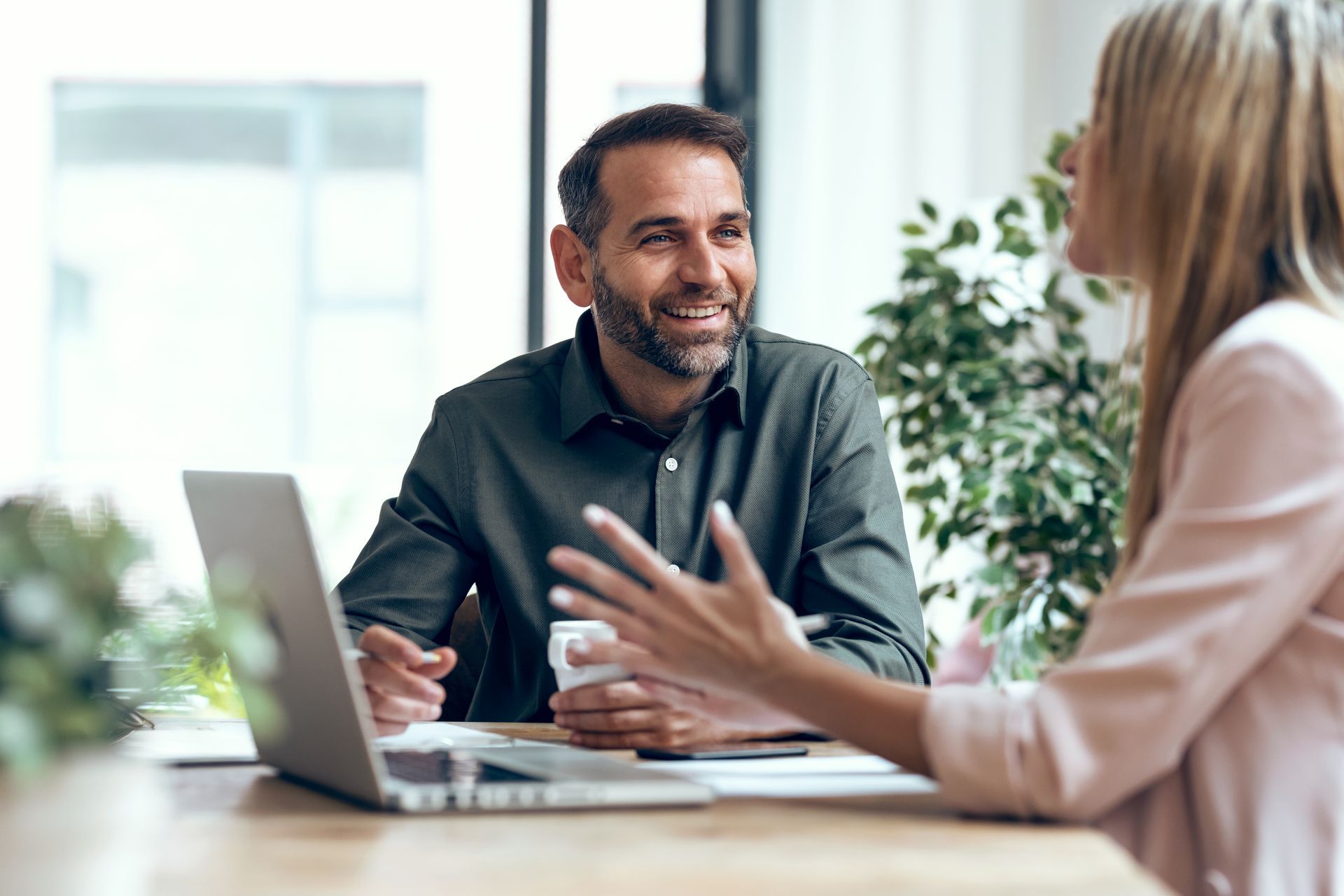 Two colleagues talking at a desk with a laptop in a bright office, gesturing during a meeting