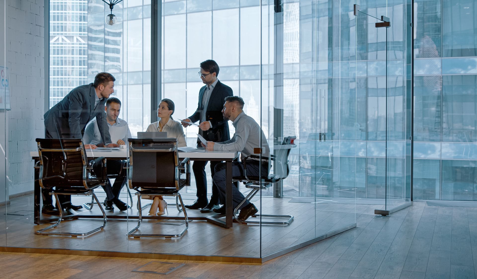 Business meeting in a glass conference room, with five people gathered around a table and laptop.
