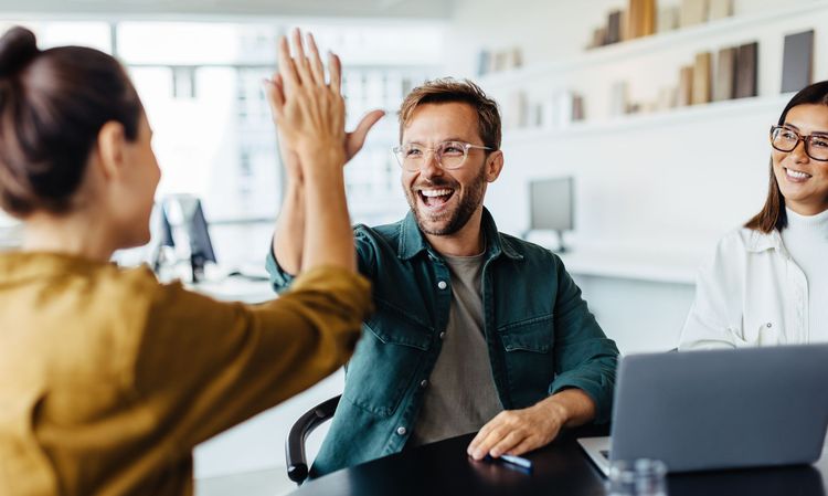 Colleagues in a bright office high-five during a meeting, with a laptop on the table.