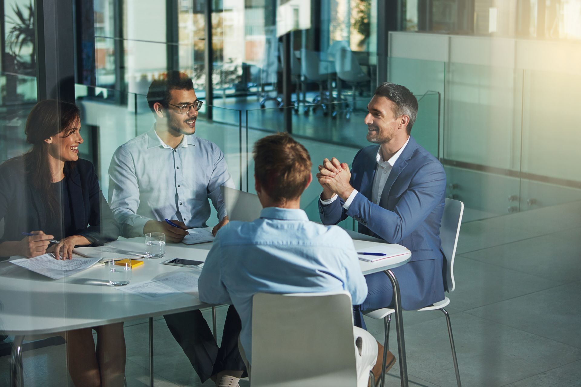 Four coworkers in a glass office meeting room, discussing around a table with papers.