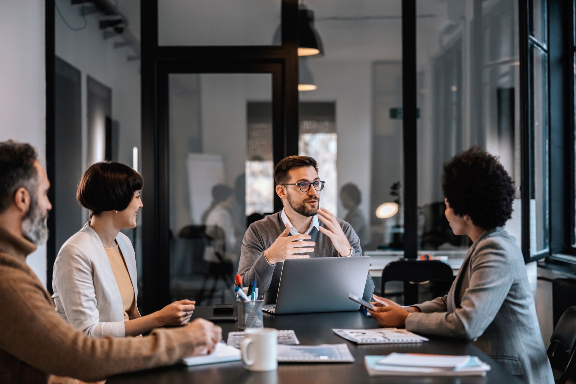 Team meeting in a modern conference room, with one person presenting and others listening around a laptop.