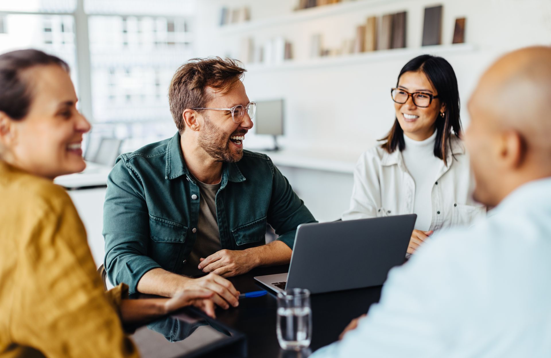 Four coworkers in a bright office meeting around a laptop, smiling and talking
