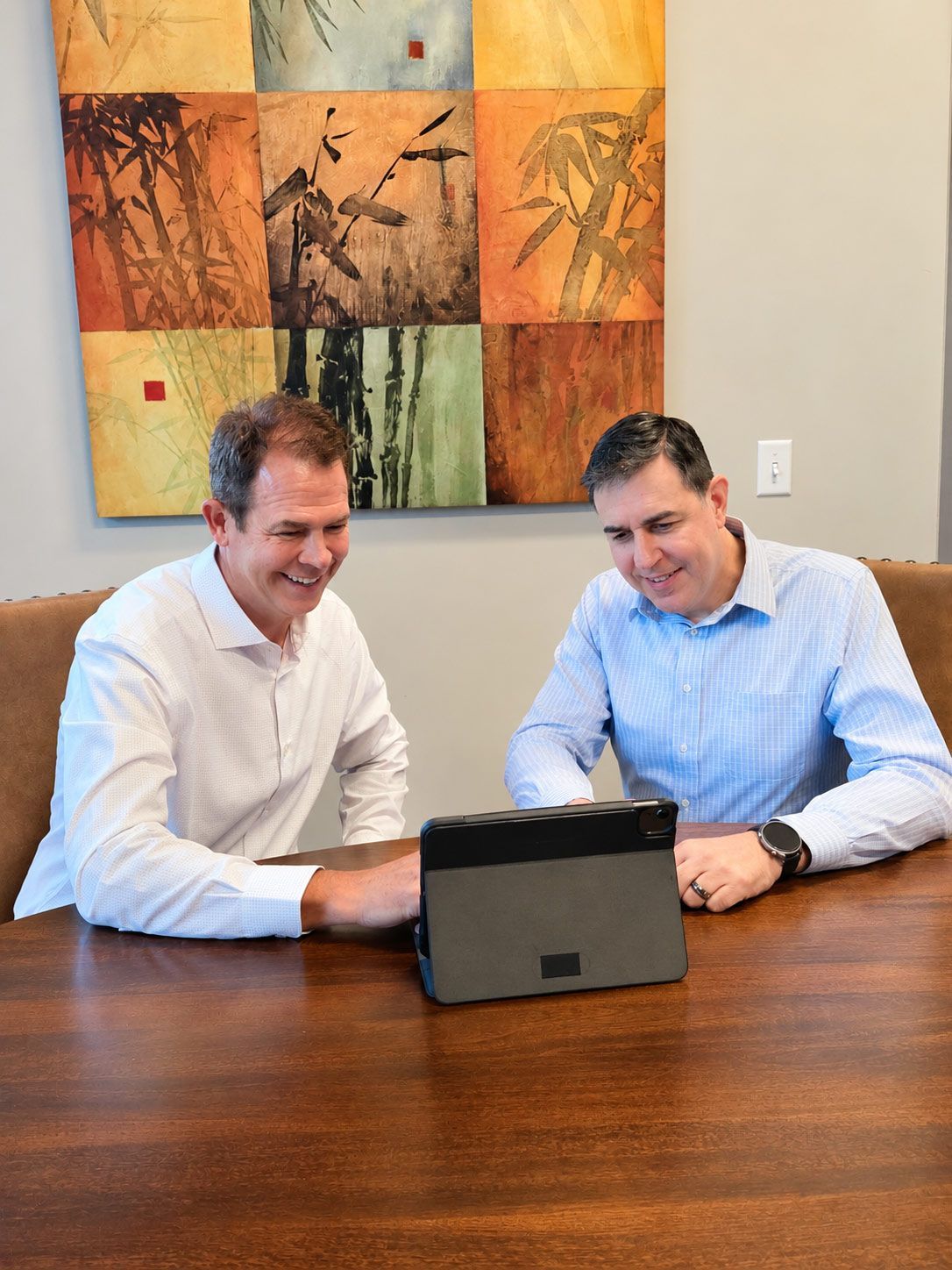 Two men in shirts leaning over a laptop at a conference table, smiling in a meeting room.