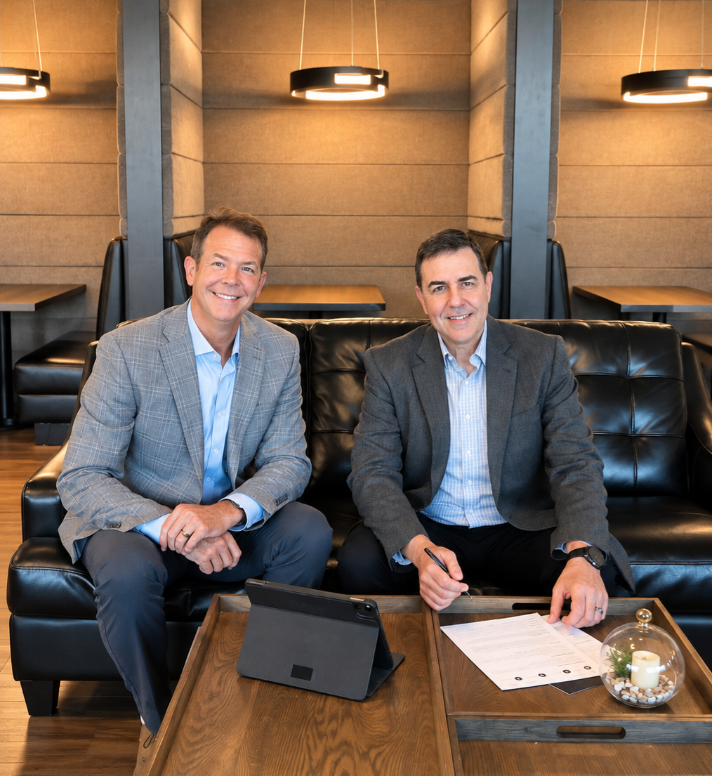 Two men in blazers standing in a bright office lobby beside a plant and lamp.