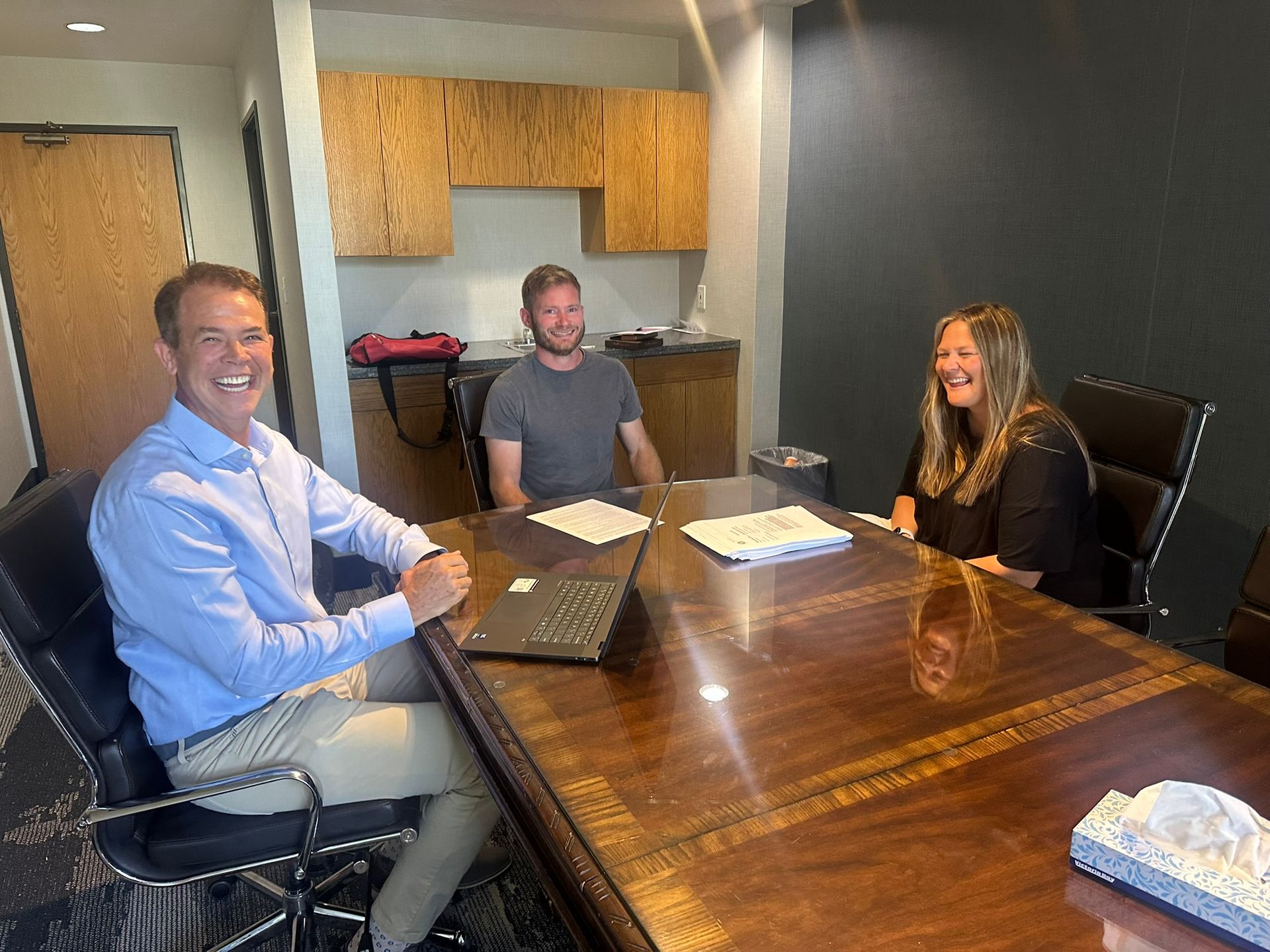 Three people seated around a conference table in a small meeting room, smiling at the camera.