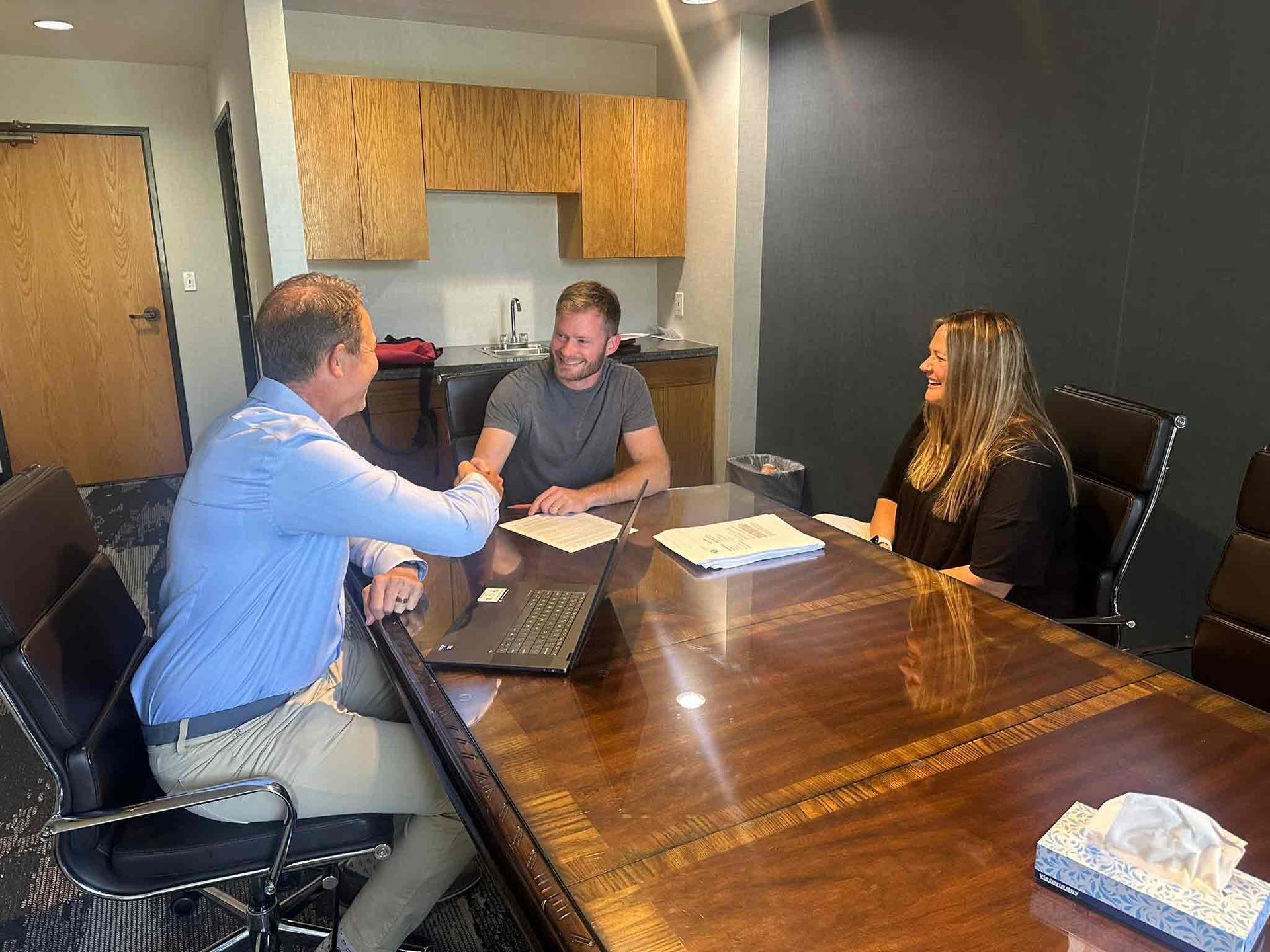 Three people in a conference room discussing documents around a wooden table with a laptop.