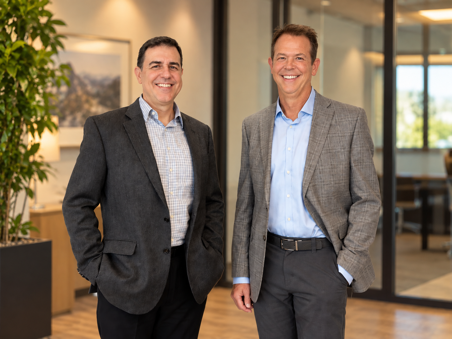 Two men in business attire standing in a modern office lobby, smiling at the camera.