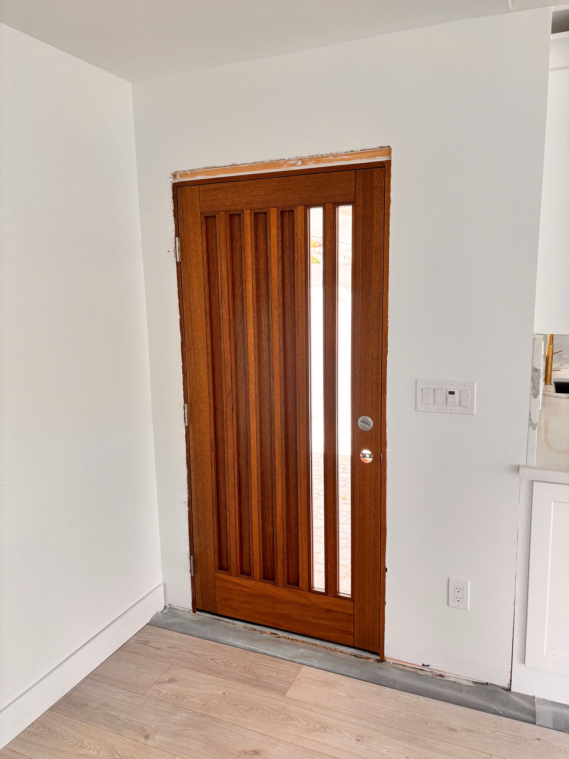 Wooden front door with narrow vertical glass panels in a white room.