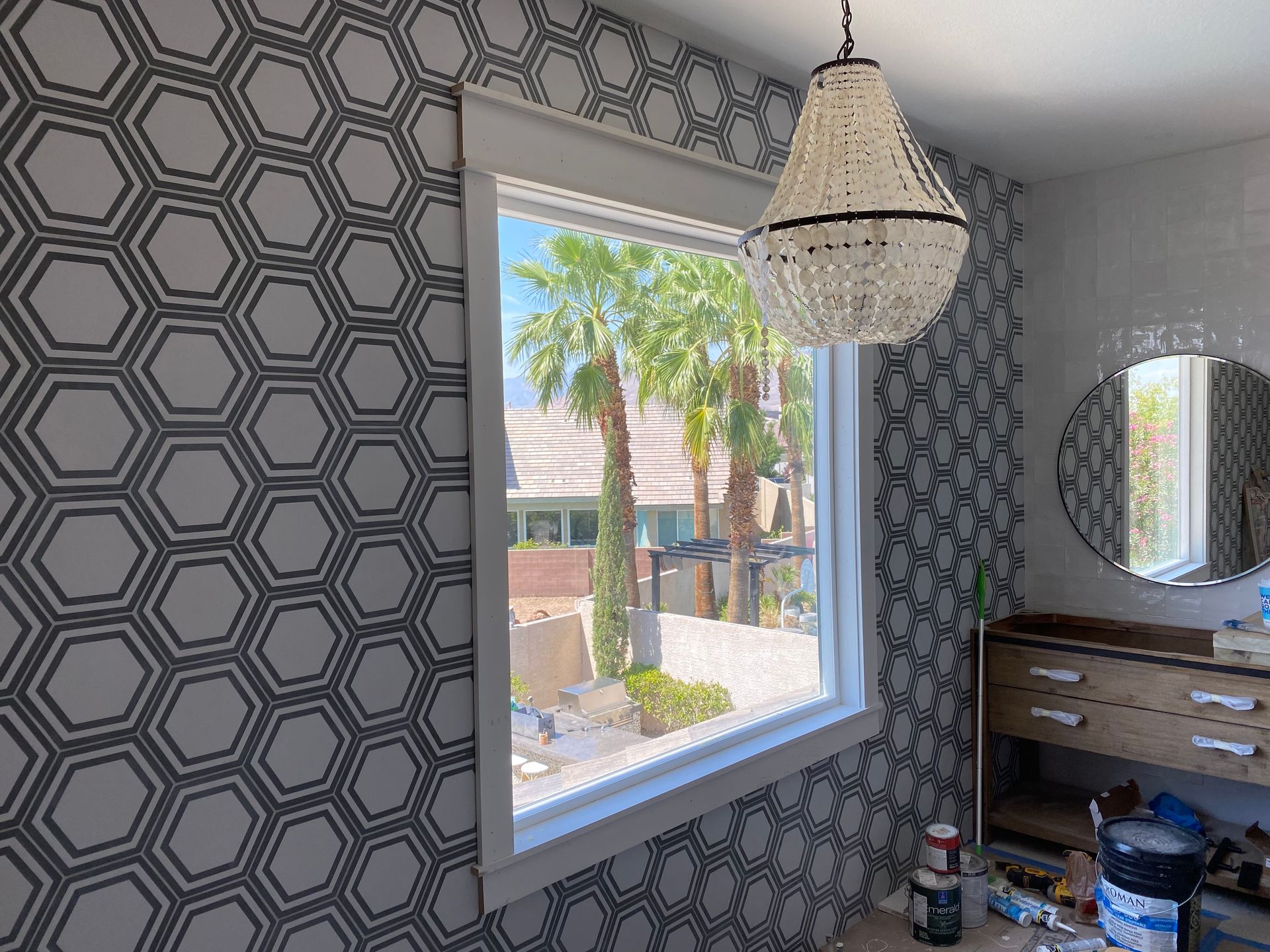 Patterned bathroom with window view, hanging light, round mirror, and wooden vanity cluttered with items