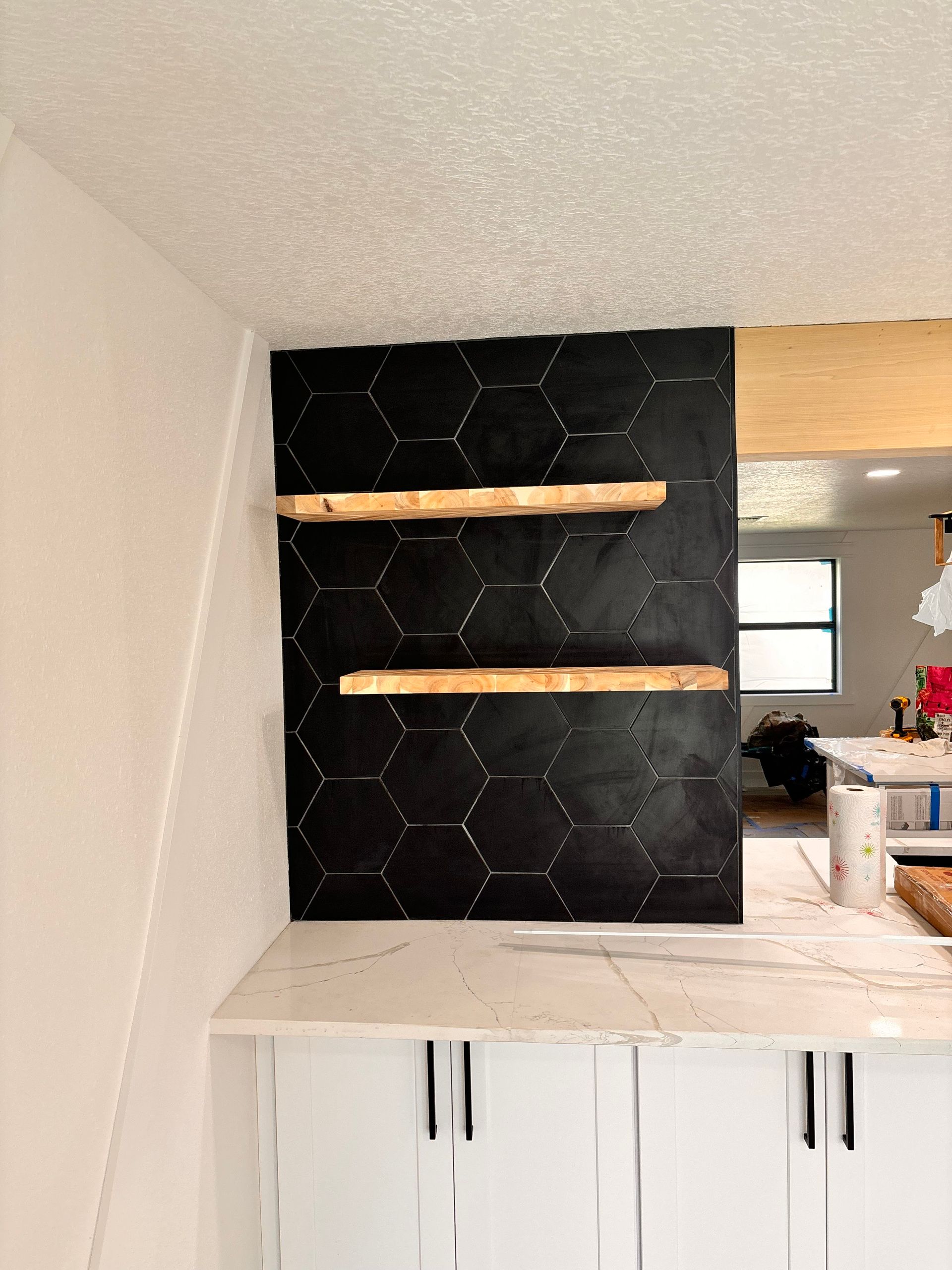 Kitchen nook with black tiled accent wall, two wood shelves, and white cabinets under a counter.