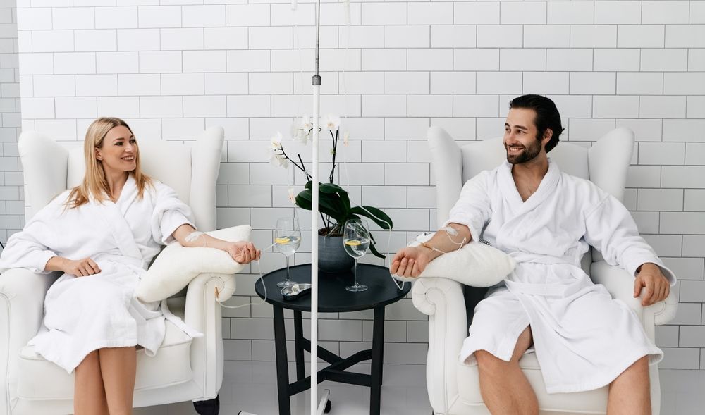 Two people in white robes chatting in a bright spa lounge with a small table and flowers.