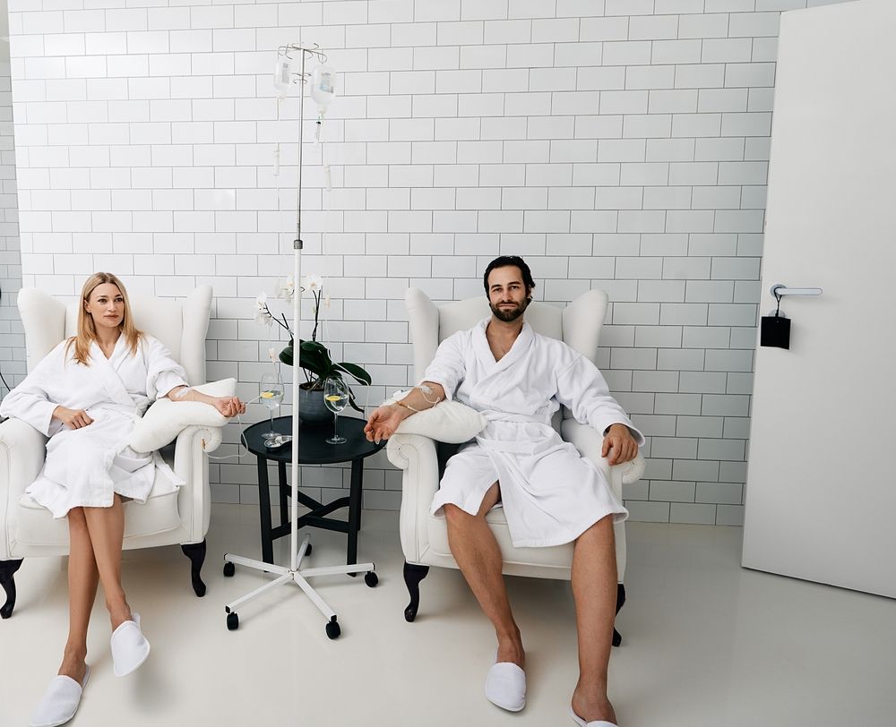Two people in white robes sit in a bright spa room, with a small table and a plant between them.