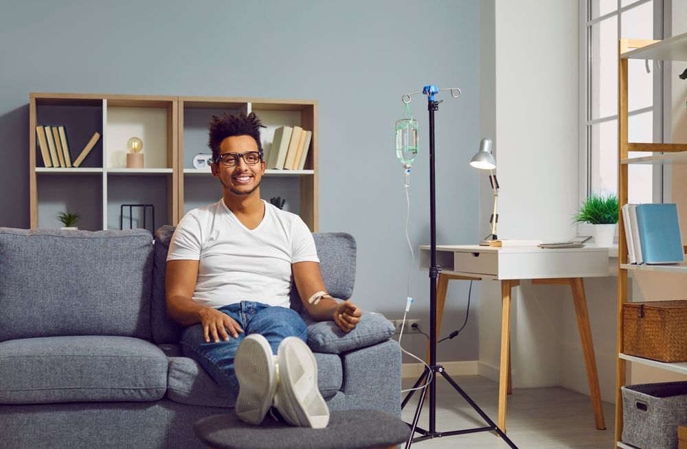 Smiling man sitting on a gray couch in a bright living room with a white desk and shelves behind him
