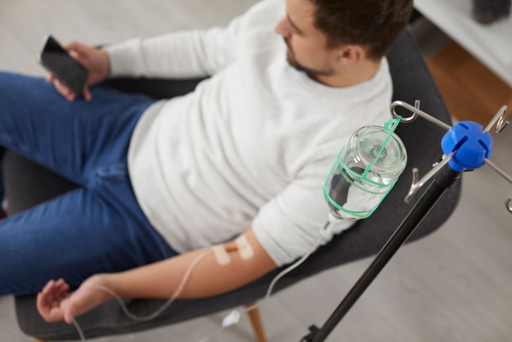 Person reclining in a chair with an IV drip and holding a phone in a home setting
