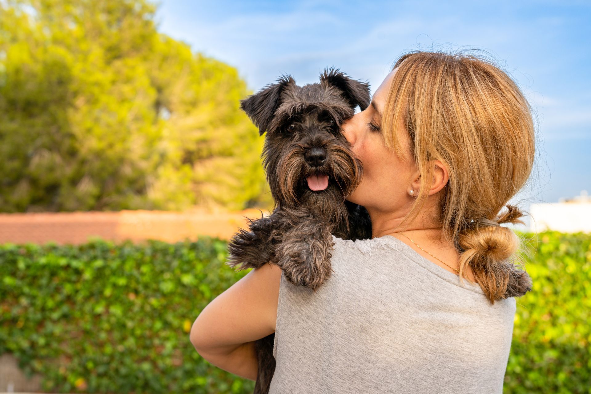 Woman holding a small black dog outdoors, both facing the camera with greenery in the background
