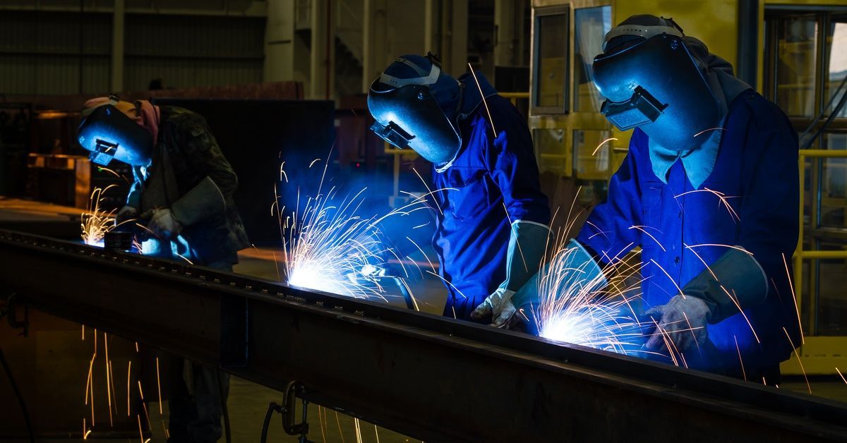 A row of workers wearing face masks and blue coveralls. They use welding tools to manufacture components in a factory.