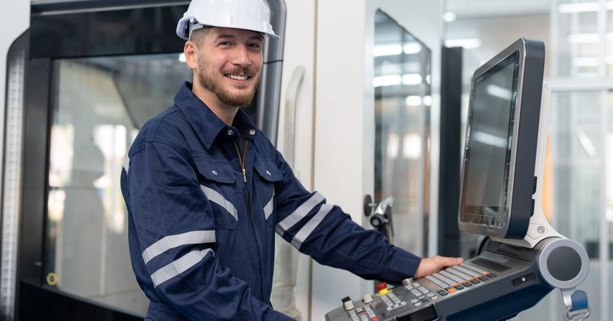 A young man wearing a blue jumpsuit and white hard hat stands at a control panel, operating a CNC machine.