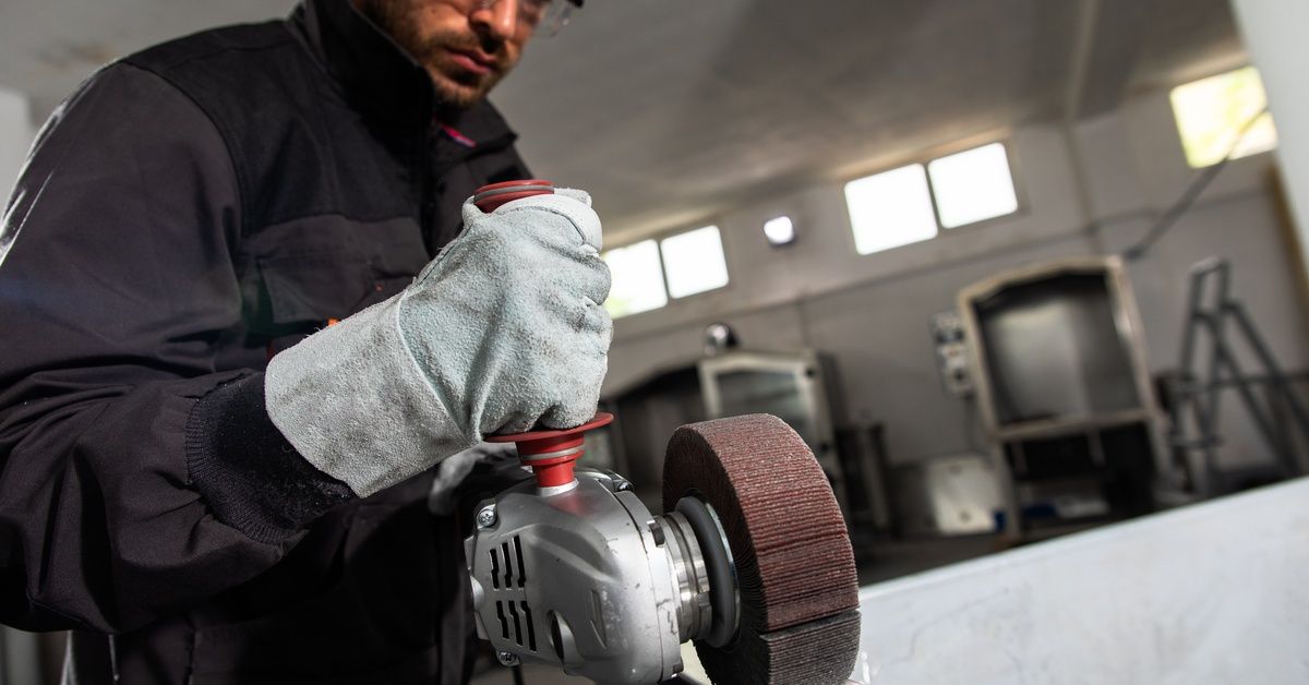 An industrial worker wearing a jumpsuit and goggles uses an angle grinder with a polishing attachment on stainless steel.