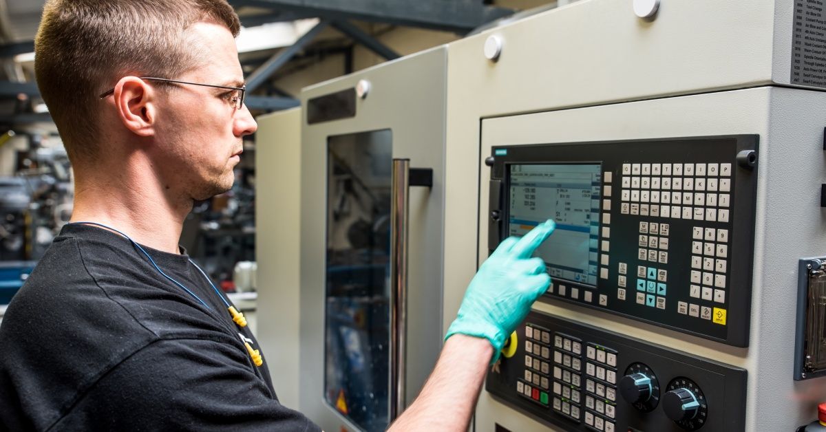 A technician wearing latex gloves taps on a touch screen on a control panel to program a CNC machine.