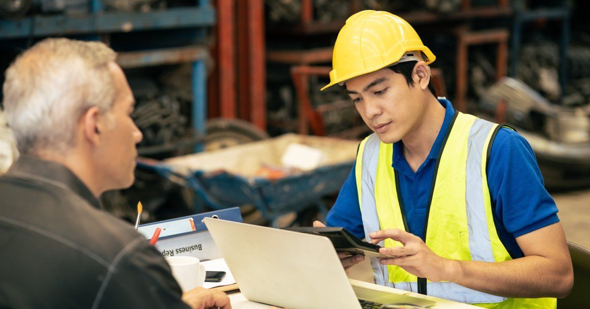 An engineer, wearing a hard hat and safety vest, sits across from an older man at a table, using a calculator.
