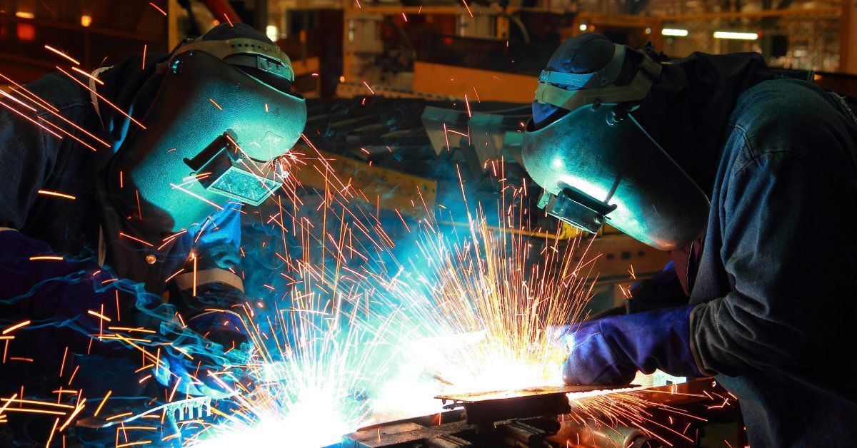 Two welders wearing helmets and protective gear work on an automotive part, creating bright sparks.