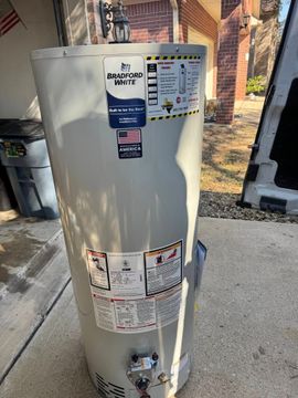 Beige water heater beside a house with warning labels and valves visible in a driveway or garage area