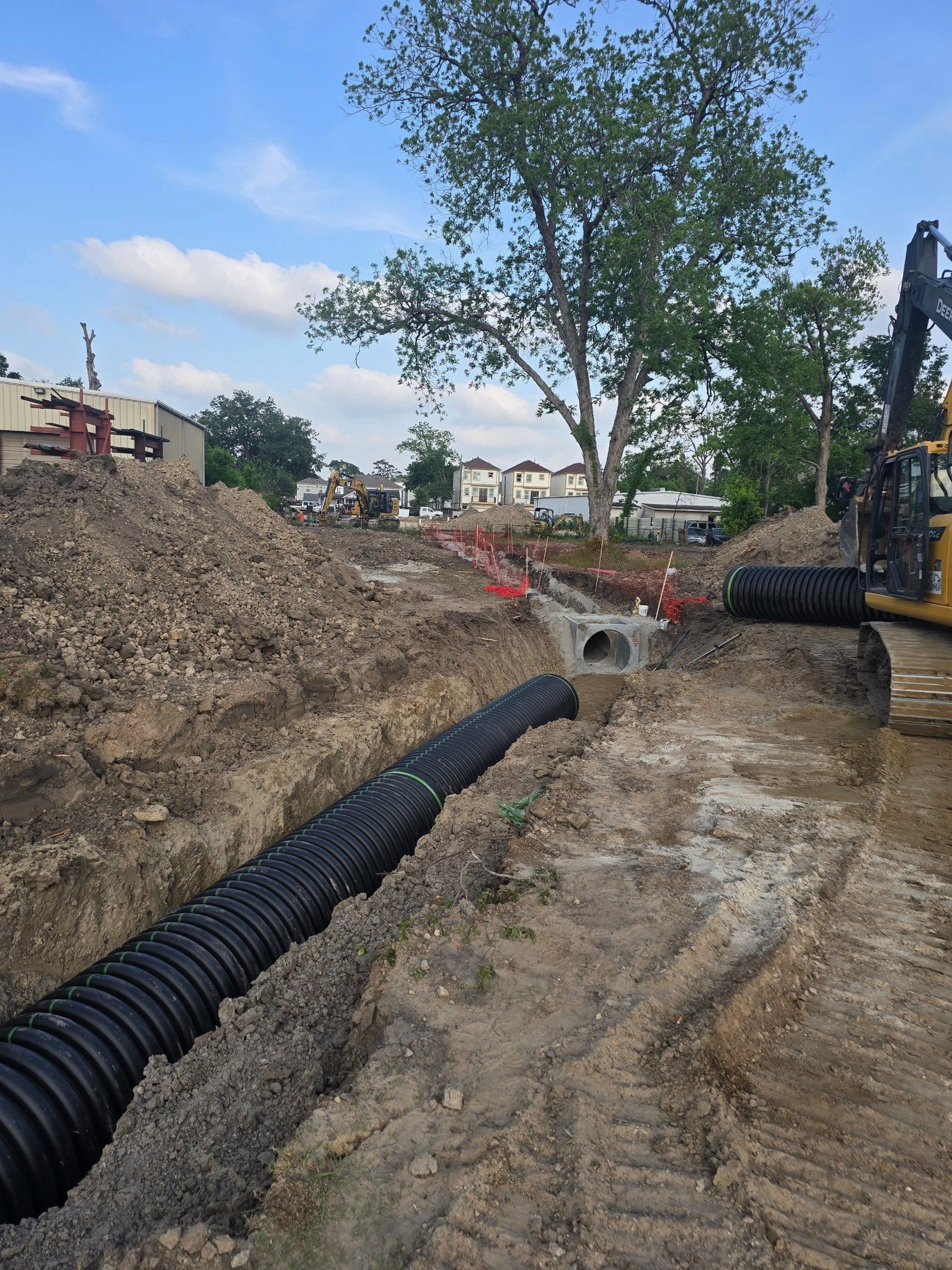 Construction trench with black corrugated pipe laid in muddy ground beside dirt piles and a tree.