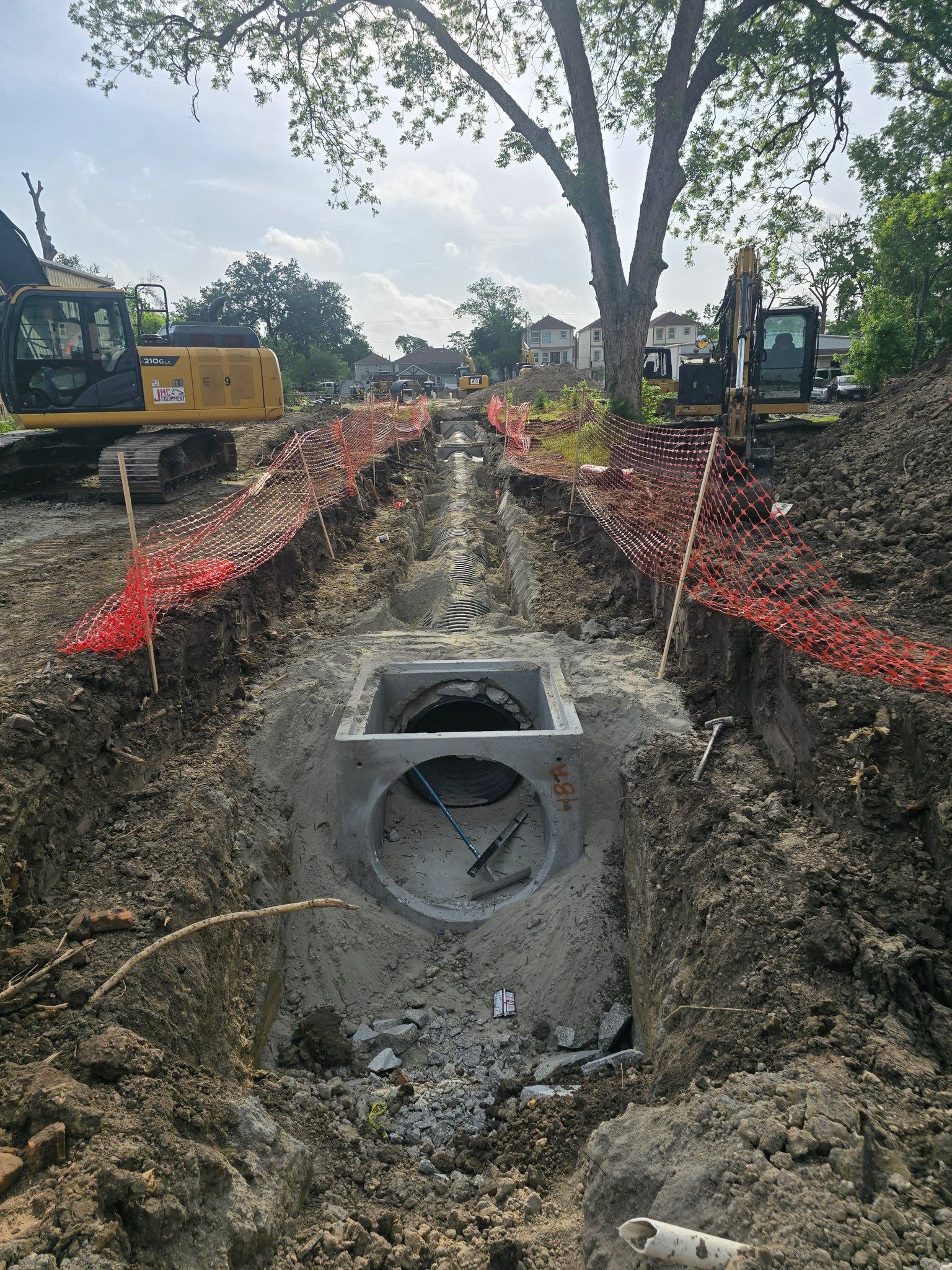 Excavation site with a large concrete pipe in a trench, lined with orange safety fencing and heavy machinery.
