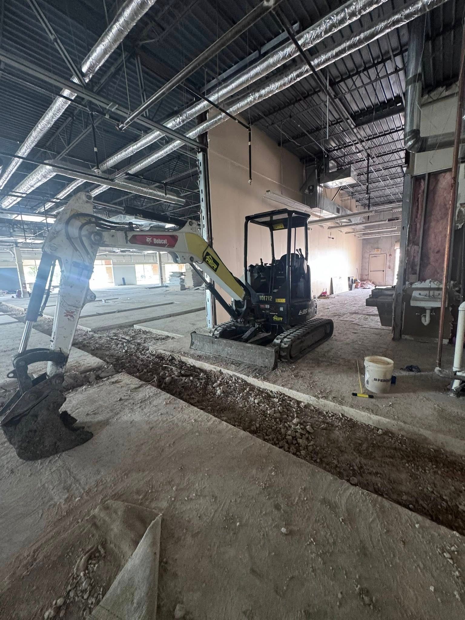 Construction site interior with excavator, piles of dirt, exposed ceiling beams, and workers in the background