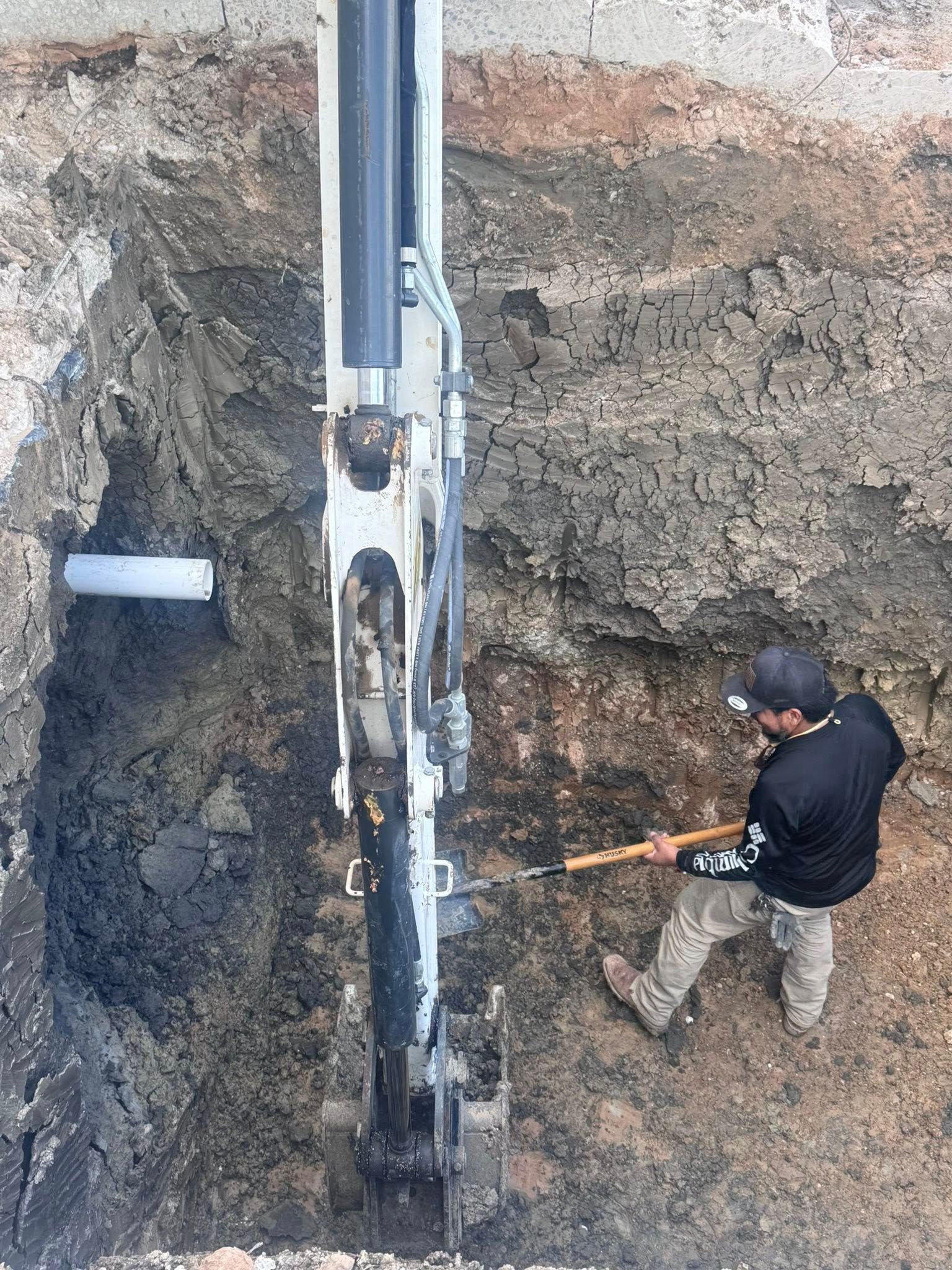 Worker operating drilling equipment in a muddy excavation pit beside a white pipe.