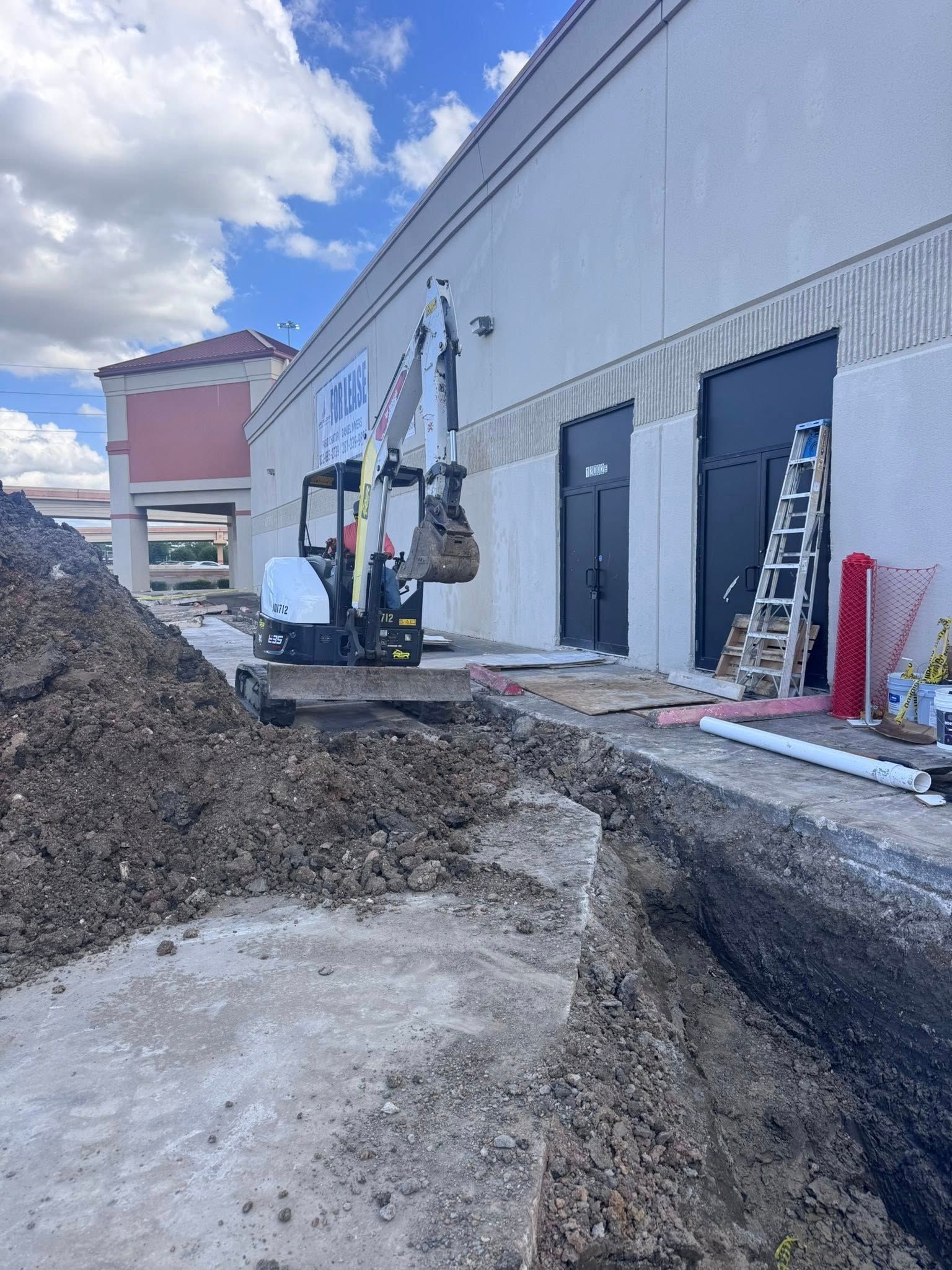 Construction site beside a gray warehouse, with an excavator digging a trench and dirt piled in the foreground.
