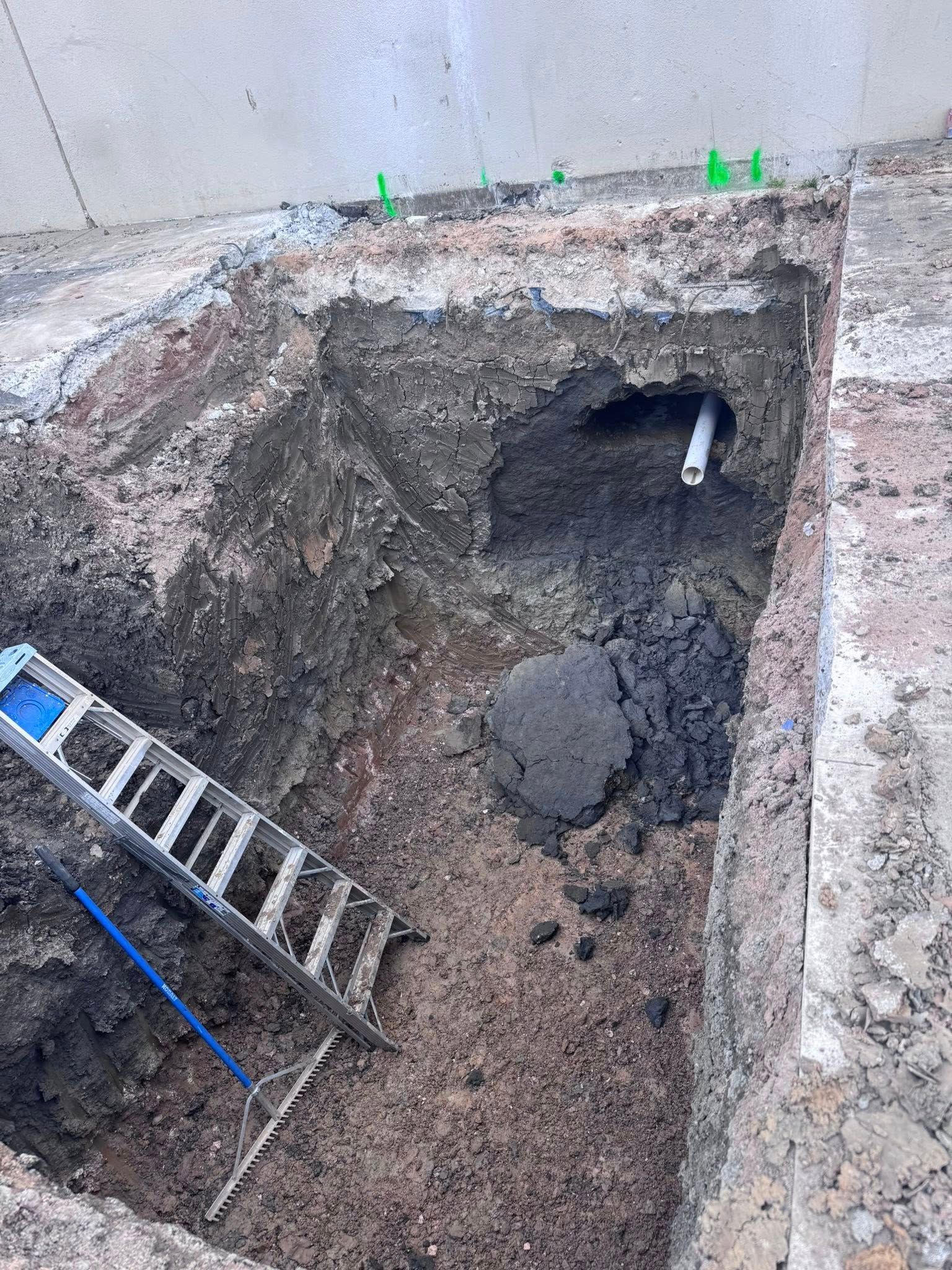 Large dug-out excavation pit beside a wall, with a ladder leaning into it and wet soil inside.