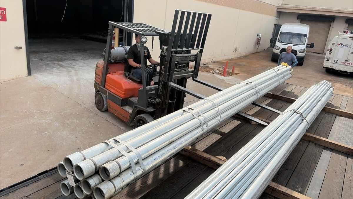 Forklift carrying long silver metal pipes in an industrial yard with more pipes stacked nearby.
