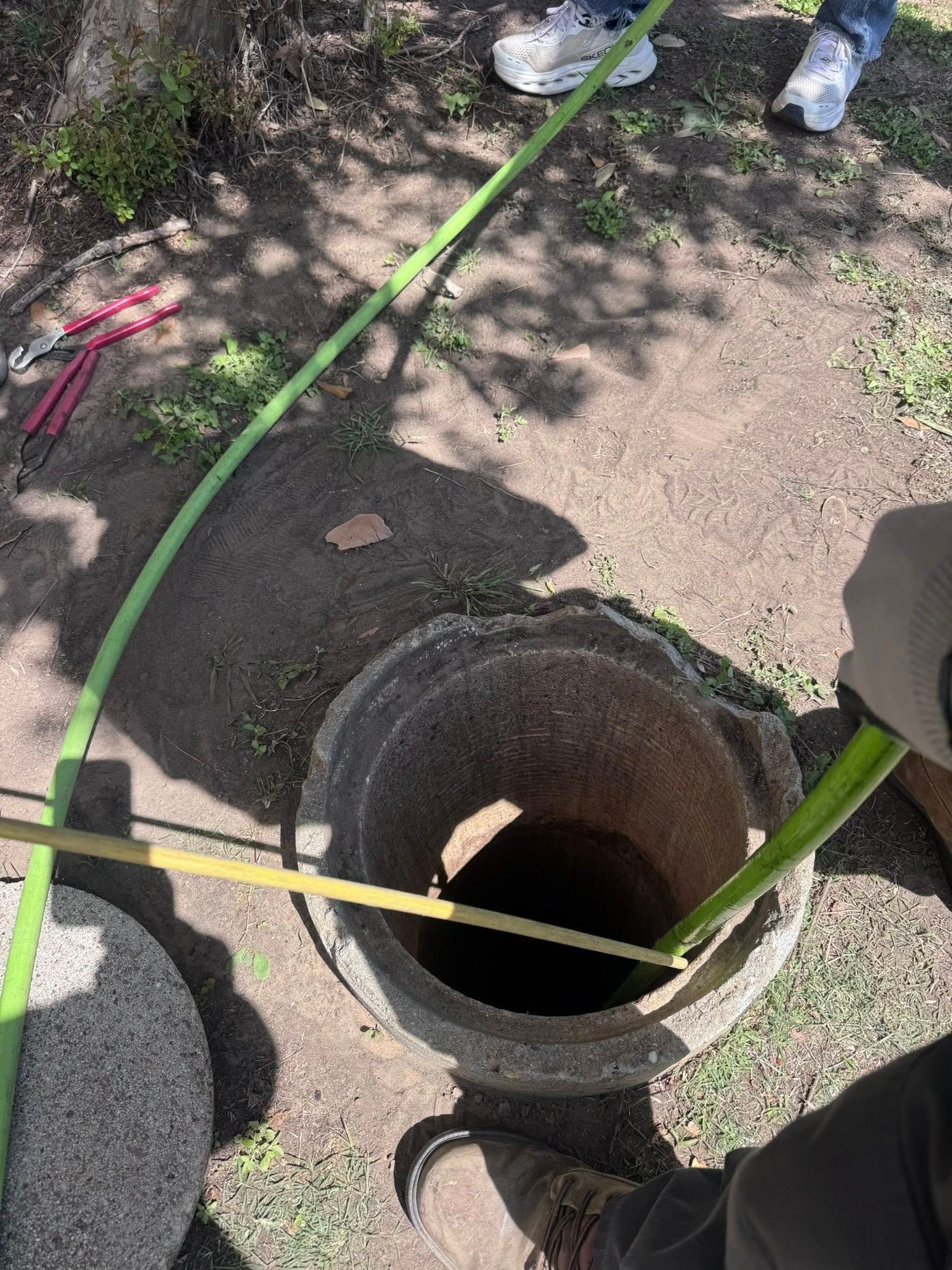 Open manhole in dirt, with green hose and yellow pole around it near workers’ feet.