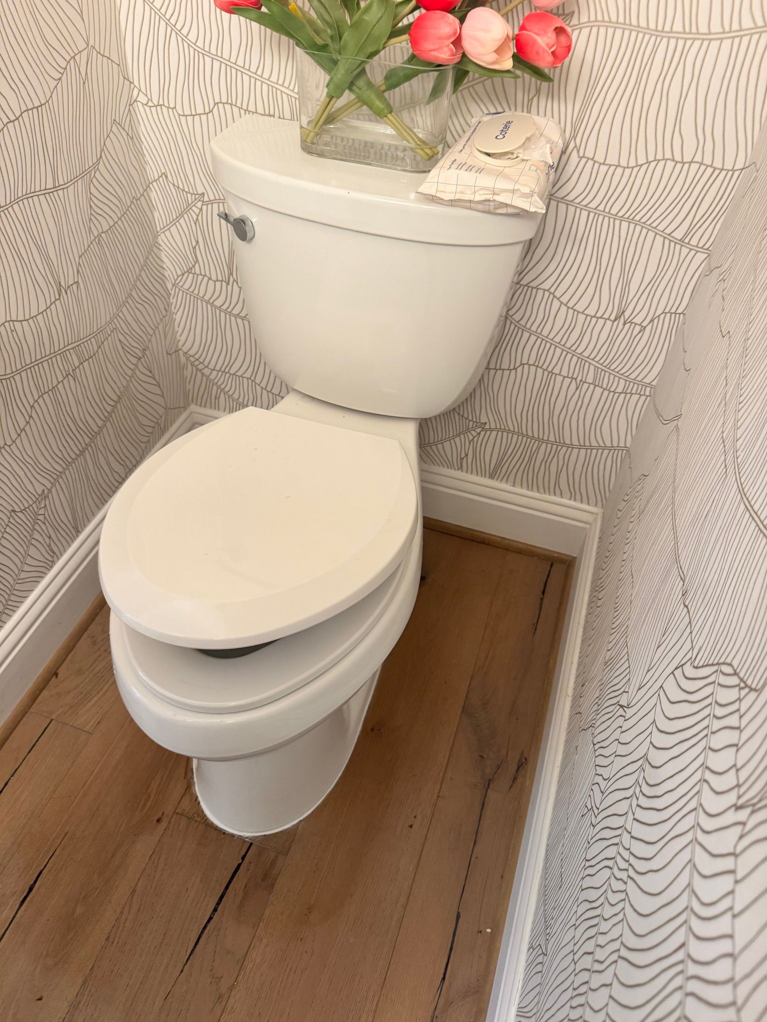 White toilet in a small bathroom with patterned wallpaper and wood-look flooring, topped with a flower arrangement