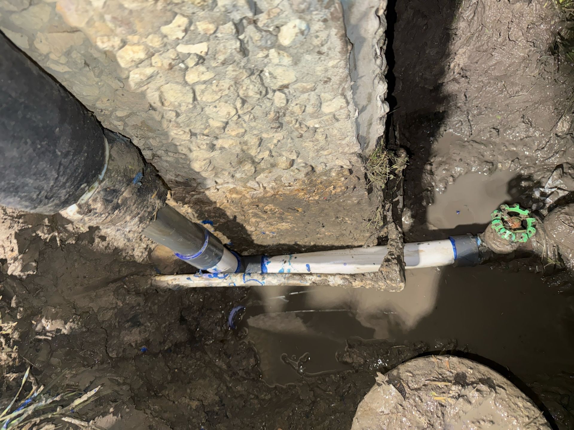 Exposed pipes and a muddy trench beside a concrete wall, with standing water and a valve at right.