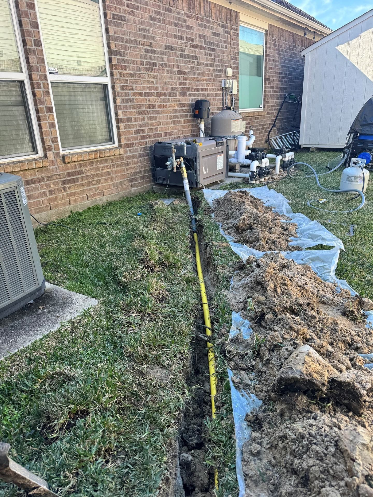 Trenched yard beside brick house, with exposed pipe line, dirt piles, and yellow tape marking the work area