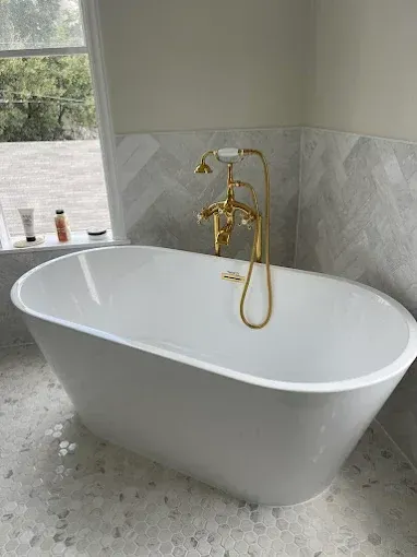 Freestanding white bathtub with gold faucet in a tiled bathroom by a window
