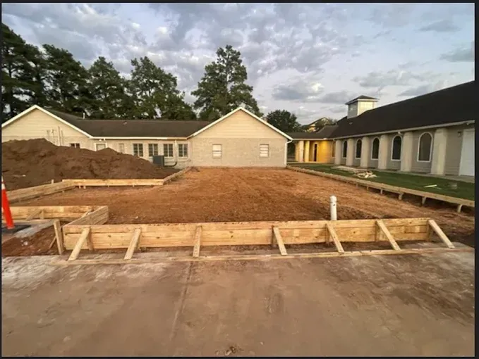 Construction site with fresh dirt and wooden forms in front of beige buildings under a cloudy sky
