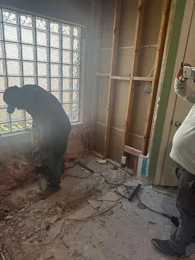 Workers clearing debris in a room with a glass-block window and exposed wall framing.